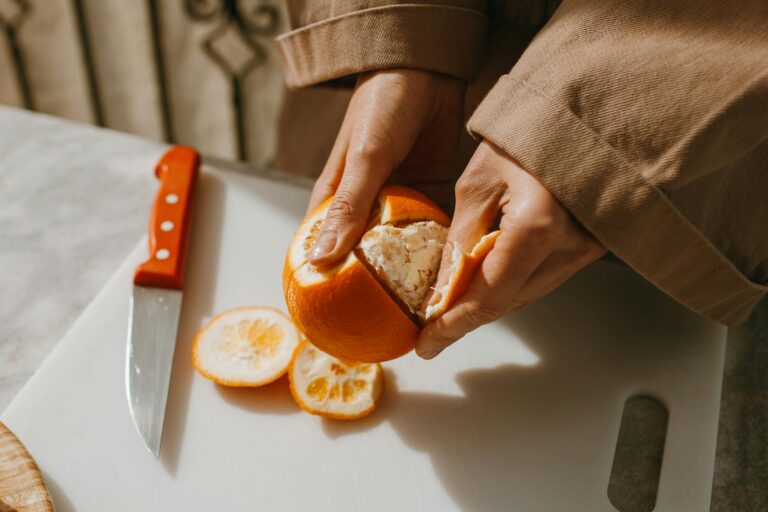 Close-up image of hands peeling a fresh orange, highlighting the texture and vibrant color on a cutting board.