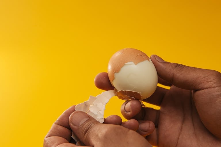 Close-up of hands peeling a boiled egg against a bright yellow backdrop, showcasing the texture.