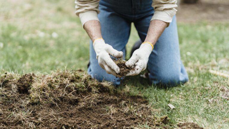 A person in jeans and gloves kneeling on grass tending to soil in a garden setting.