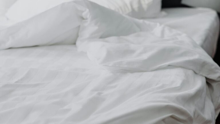 A close-up of a cozy white linen bed with rumpled bedding in a modern bedroom setting.