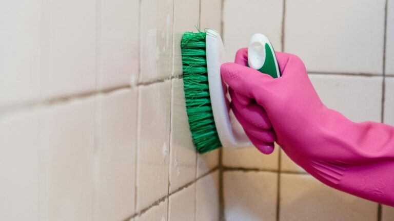 A person scrubs bathroom tiles wearing a pink glove, emphasizing cleanliness.