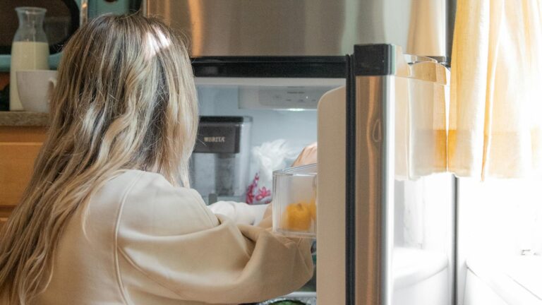 A woman reaches inside a refrigerator in a sunlit kitchen, arranging groceries.