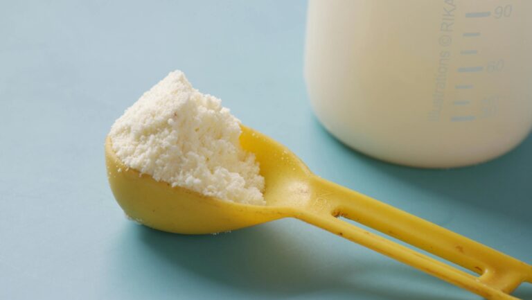 A yellow spoon filled with powdered milk, next to a measuring bottle, on a blue background.