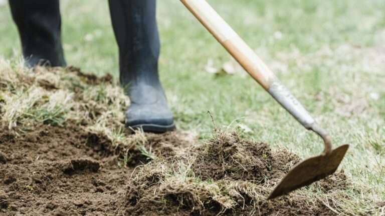 Crop unrecognizable farmer in dirty rubber boots standing and loosening dry soil in countryside in daylight