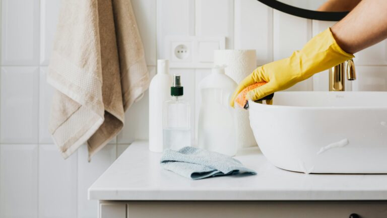 Person cleaning a bathroom sink with yellow gloves and cleaning products on a countertop.