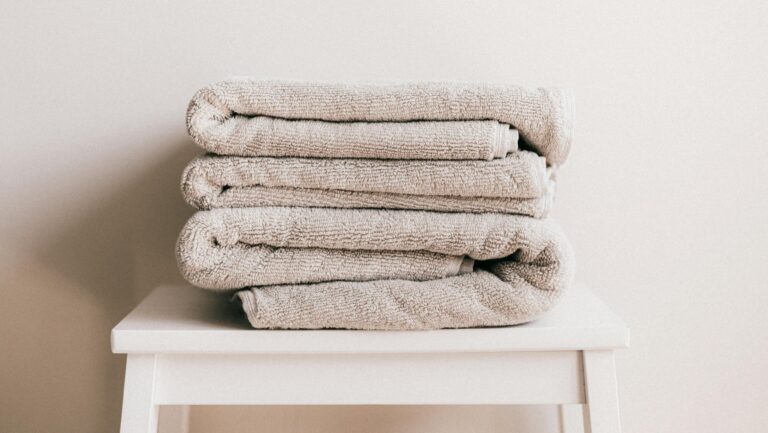 Stack of soft clean light beige folded towels placed on white minimalist stool against beige wall in daylight