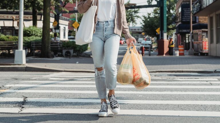 Woman wearing face mask and carrying groceries on zebra crossing in urban area with train overhead.